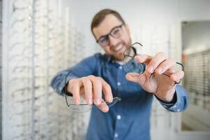 Satisfied Customer. View of happy young male client wearing new glasses, standing near rack and showcase with eyewear. Smiling man trying on spectacles photo