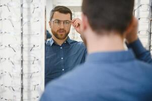 In Optics Shop. Portrait of male client holding and wearing different spectacles, choosing and trying on new glasses at optical store. Man picking frame for vision correction, closeup. photo