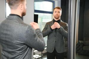 moderno joven hermoso empresario vestido en clásico traje ajustando un Corbata en frente de el espejo mientras en pie en el traje tienda foto