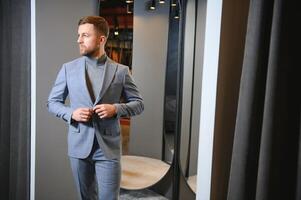 Modern young handsome businessman dressed in classical suit adjusting a tie in front of the mirror while standing in the suit shop photo