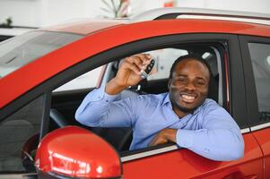 contento coche comprador, nuevo coche propietario concepto. retrato de emocionado joven africano americano chico en concesión sala de exposición foto