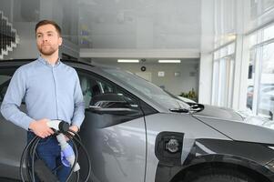 barba hombre molesto un nuevo cargando cable con un coche cargando estación a el motor concesión. concepto de comprando eléctrico vehículo. inteligente ecológico vivo foto