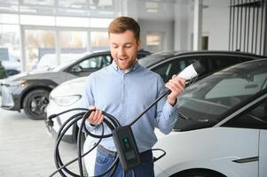 Handsome business man holding charging cable for electric car. Caucasian male stands near electric auto in dealership photo