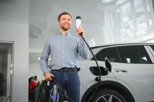 Handsome business man holding charging cable for electric car. Caucasian male stands near electric auto in dealership photo