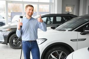 Handsome business man holding charging cable for electric car. Caucasian male stands near electric auto in dealership photo