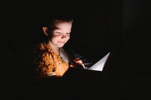 Child reading book in bed. Kids read at night. Little boy with fairy tale books in bedroom . Education for young children. Bedtime story in the evening. Cute kid under blanket in dark room with lamp photo