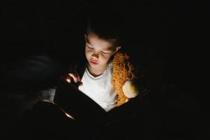 retrato de linda pequeño chico leyendo en cama con Linterna en oscuro habitación, disfrutando cuentos de hadas. foto