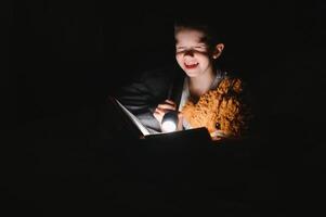 un chico de 5-6 años antiguo es leyendo un libro en el noche en el oscuro debajo un cobija con un juguete oso. foto