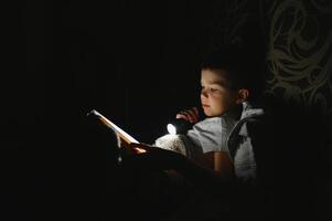 niño leyendo libro en cama. niños leer a noche. pequeño chico con hada cuento libros en dormitorio . educación para joven niños. hora de acostarse historia en el noche. linda niño debajo cobija en oscuro habitación con lámpara foto