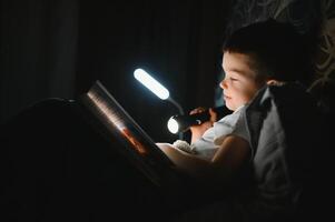 Reading book and using flashlight. Young boy in casual clothes lying down near tent at evening time. photo