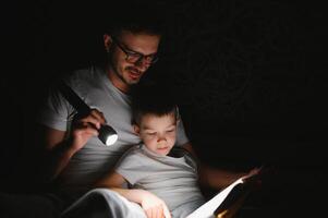 Happy family reading bedtime story under blanket in evening. Father and son spend time together. Father's Day photo