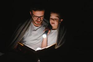 Happy family reading bedtime story under blanket in evening. Father and son spend time together. Father's Day photo