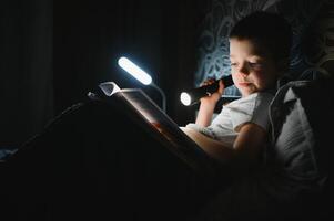 Reading book and using flashlight. Young boy in casual clothes lying down near tent at evening time. photo