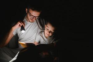 Father and son with flashlight reading book under blanket at home. photo