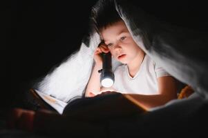 Reading book and using flashlight. Young boy in casual clothes lying down near tent at evening time. photo