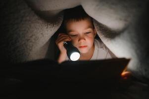 Reading book and using flashlight. Young boy in casual clothes lying down near tent at evening time photo