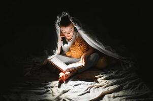 un chico de 5-6 años antiguo es leyendo un libro en el noche en el oscuro debajo un cobija con un juguete oso. foto