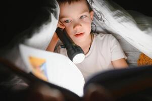 niño leyendo libro en cama. niños leer a noche. pequeño chico con hada cuento libros en dormitorio . educación para joven niños. hora de acostarse historia en el noche. linda niño debajo cobija en oscuro habitación con lámpara foto