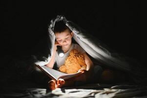 chico con Linterna leyendo libro debajo cobija a hogar. foto