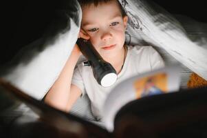 Reading book and using flashlight. Young boy in casual clothes lying down near tent at evening time photo