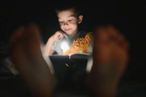 Reading book and using flashlight. Young boy in casual clothes lying down near tent at evening time photo