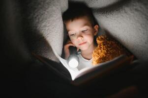 Reading book and using flashlight. Young boy in casual clothes lying down near tent at evening time photo