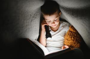 chico con Linterna leyendo libro debajo cobija a hogar. foto