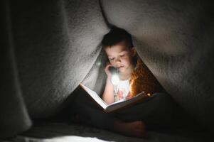 niño leyendo libro en cama. niños leer a noche. pequeño chico con hada cuento libros en dormitorio . educación para joven niños. hora de acostarse historia en el noche. linda niño debajo cobija en oscuro habitación con lámpara foto