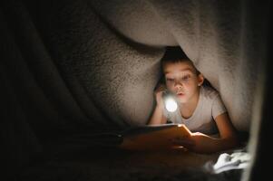 Reading book and using flashlight. Young boy in casual clothes lying down near tent at evening time. photo