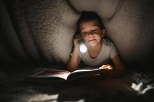 retrato de linda pequeño chico leyendo en cama con Linterna en oscuro habitación, disfrutando cuentos de hadas foto