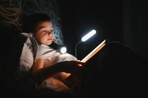 niño leyendo libro en cama. niños leer a noche. pequeño chico con hada cuento libros en dormitorio . educación para joven niños. hora de acostarse historia en el noche. linda niño debajo cobija en oscuro habitación con lámpara foto