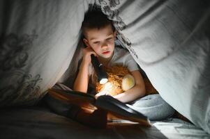 chico con Linterna leyendo libro debajo cobija a hogar foto
