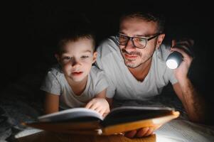 Father and son with flashlight reading book under blanket at home. photo