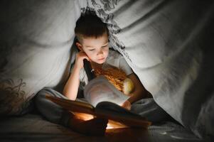 chico con Linterna leyendo libro debajo cobija a hogar. foto