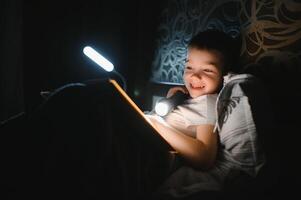 Reading book and using flashlight. Young boy in casual clothes lying down near tent at evening time photo