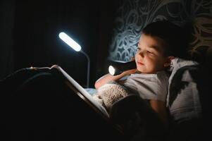 Reading book and using flashlight. Young boy in casual clothes lying down near tent at evening time photo