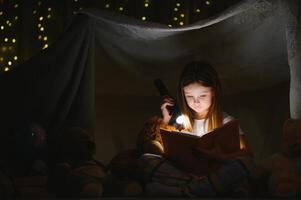 niño niña leyendo con libro y Linterna y osito de peluche oso en carpa. antes de yendo a cama. foto