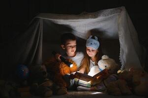 Siblings sit in a hut of chairs and blankets. Brother and sister reading book with flashlight at home. photo