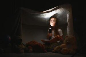 pequeño niña son leyendo un libro con linternas en carpa. contento niña jugando a hogar. gracioso encantador niño teniendo divertido en habitación foto