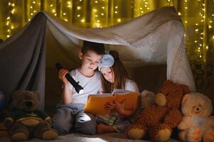 dos pequeño niño jugar a hogar en el noche a construir un cámping tienda a leer libros con un Linterna y dormir adentro. concepto de juego, magia, creatividad, alarma sistemas foto