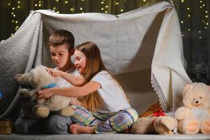 dos pequeño niño jugar a hogar en el noche a construir un cámping tienda a leer libros con un Linterna y dormir adentro. concepto de juego, magia, creatividad, alarma sistemas foto