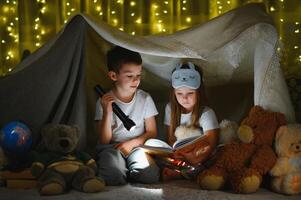 dos niños con Linterna leer un libro debajo un cobija como un carpa. foto