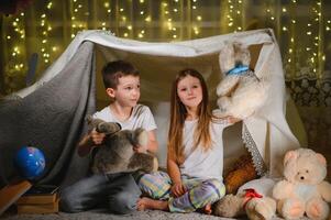 Two little children play at home in the evening to build a camping tent to read books with a flashlight and sleep inside. Concept of game, magic, creativity, alarm systems. photo