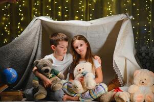 dos pequeño niños jugar a hogar en el noche a construir un cámping tienda a leer libros con un Linterna y dormir adentro. concepto de juego, magia, creatividad, alarma sistemas foto