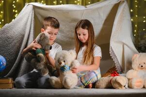 Two little child play at home in the evening to build a camping tent to read books with a flashlight and sleep inside. Concept of game, magic, creativity, alarm systems. photo