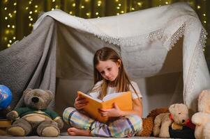 child girl reading with book and flashlight and teddy bear in tent. before going to bed. photo