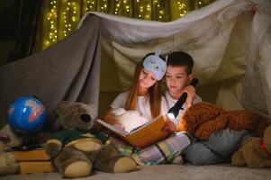 dos niños con Linterna leer un libro debajo un cobija como un carpa. foto