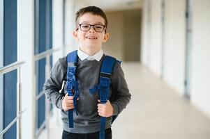 alegre sonriente pequeño chico con grande mochila teniendo divertida. colegio concepto. espalda a colegio foto