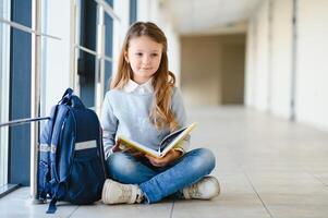 Little pretty girl sitting on the floor of the school hallway and reading a book. The concept of schooling. photo