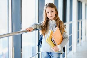 Front view of little beautiful school girl among corridor at school, holding notes at hands. Funny and happy girl smiling at camera, resting after lessons on primary school photo